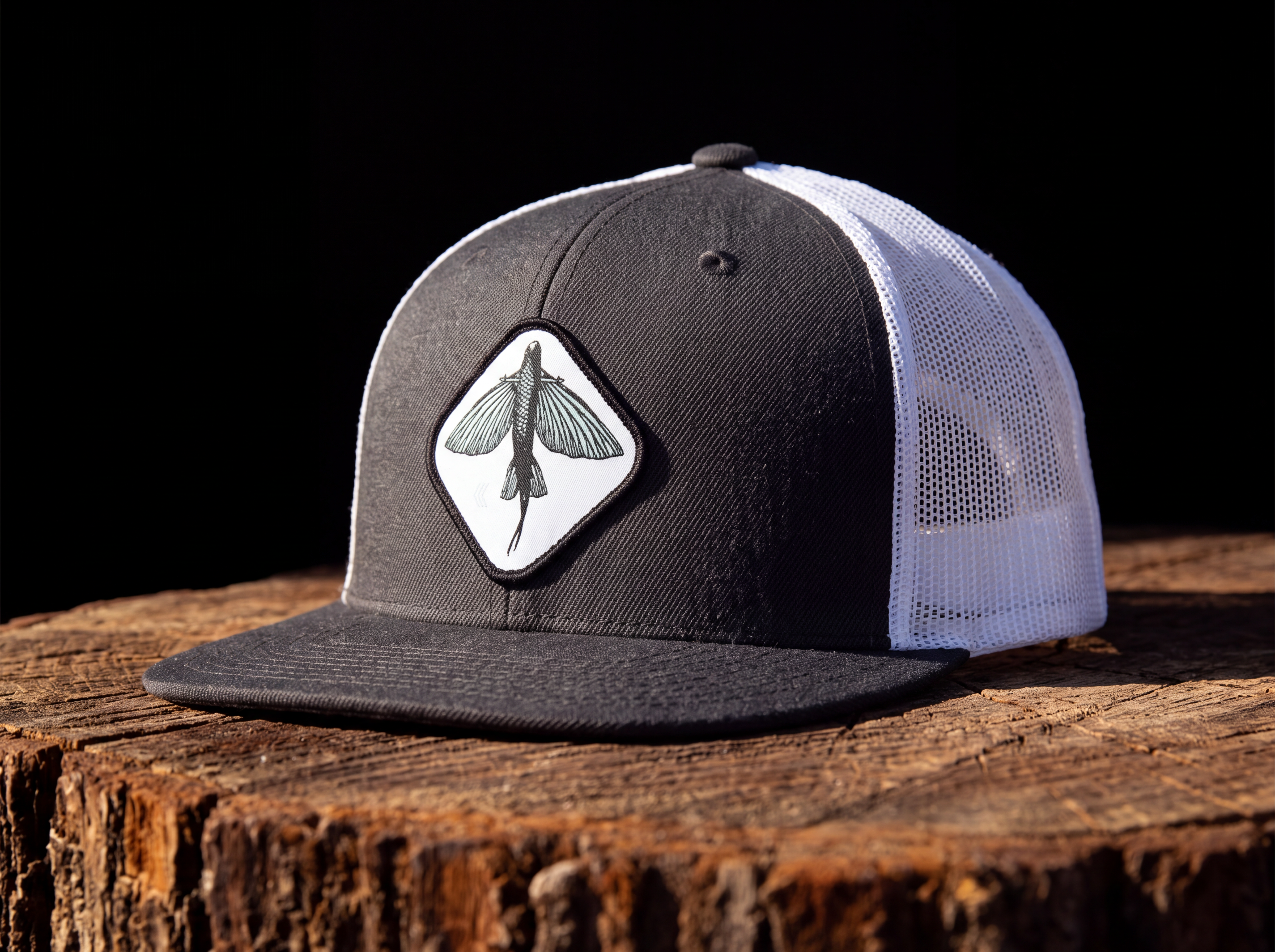 A black and white trucker cap with a white mesh back and a black flat brim, featuring a diamond-shaped patch with a black fish design on the front. The cap is displayed on a rustic wooden log against a solid black background.