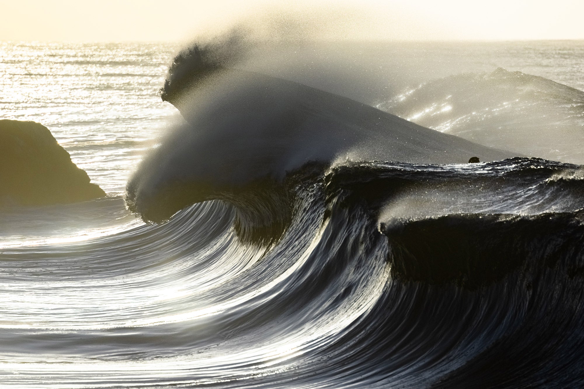 Large wave crashing against a rocky shore with sunlight reflecting off the water.