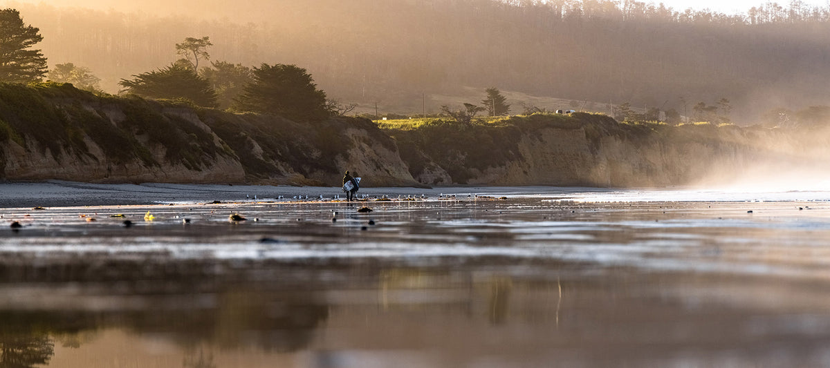 A scenic coastal landscape print showing a person walking on a wet shoreline with cliffs, trees, and misty golden light in the background.